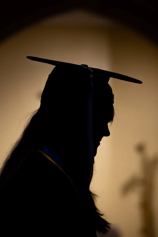 Silhouette of a graduating student wearing a cap and gown, head bowed in a dimly lit space, possibly a church, with a blurred cross visible in the background.