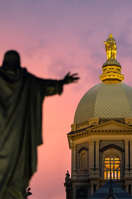 Silhouetted statue with outstretched arms against a vibrant pink and orange sunset, with the golden dome of the Main Building and its statue of Mary at the University of Notre Dame