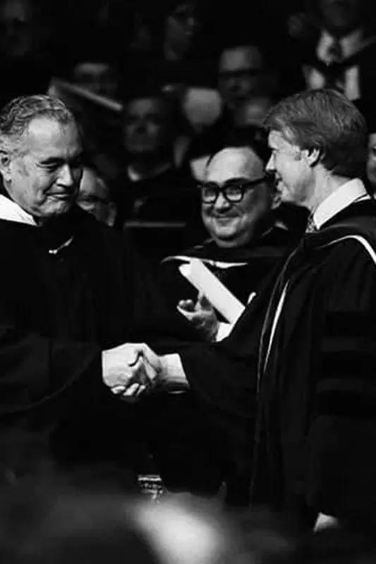 A black and white photo shows Jimmy Carter shaking hands with Father Hesburgh in academic regalia.