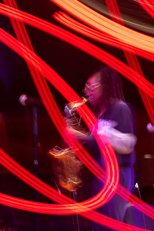 A person with dreadlocks and glasses plays a saxophone, blurred by red light streaks.