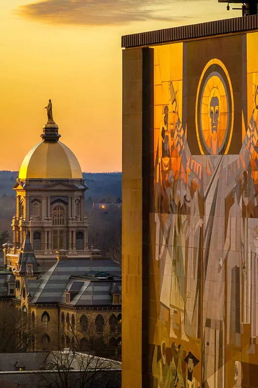The Golden Dome of the Main Building and the &ldquo;Word of Life&rdquo; mural on the Hesburgh Library at Notre Dame are bathed in warm, golden light at sunset.