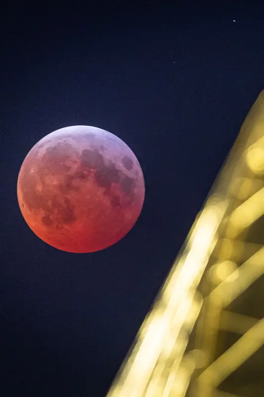 A close-up vertical shot of a reddish-orange moon in a dark sky next to a blurred golden dome.