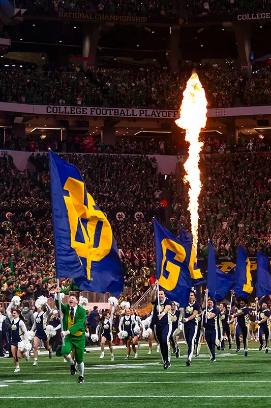 A Notre Dame leprechaun in a green suit runs onto a field with cheerleaders and flag bearers as a pyrotechnic flame shoots upwards, with a stadium crowd in the background.