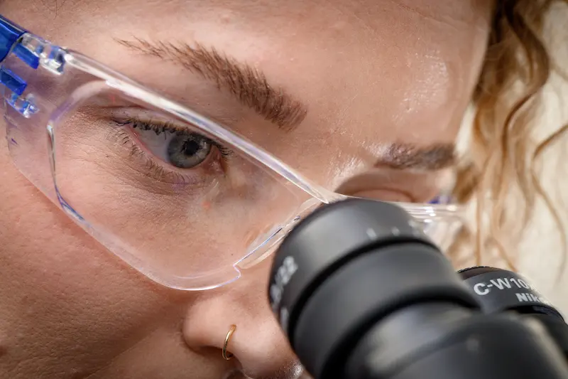 A female researcher with curly hair and clear safety glasses looks intently into a microscope.