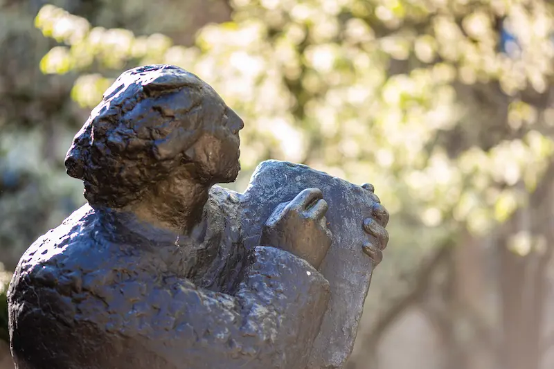 A close-up of a bronze statue of a person looking upward, holding a book or tablet, with blurred sunlight and foliage in the background.