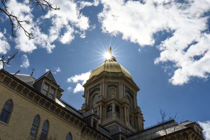 A low-angle view of the Golden Dome of the Main Building at Notre Dame, with the sunburst directly above it against a bright blue sky with clouds.