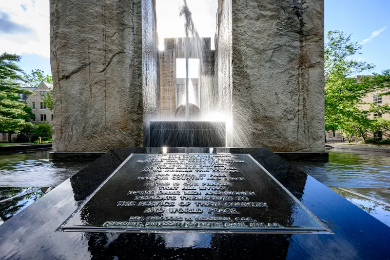 A large stone monument with water cascading down its sides has a polished black surface in the foreground with a reflective inscription and a cross visible through the water.