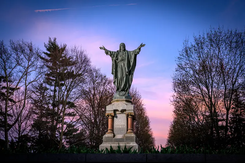 The Sacred Heart of Jesus statue at Notre Dame, with arms outstretched, is seen against a backdrop of bare trees and a vibrant pink and blue sunset sky.
