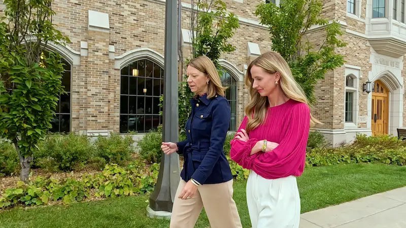 Two women walk on a sidewalk next to a brick building, one in a navy blue jacket and tan pants, the other in a bright pink blouse and white pants.