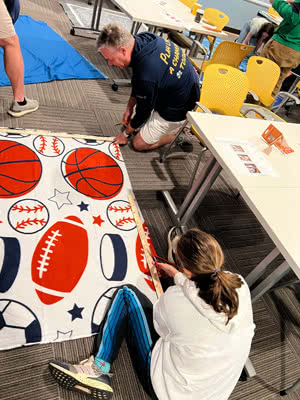 Two people sit on the floor in a classroom cutting fabric to make blankets.