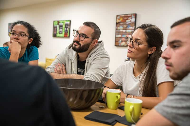 Four students sit at a table with coffee mugs and listen to other students talk.