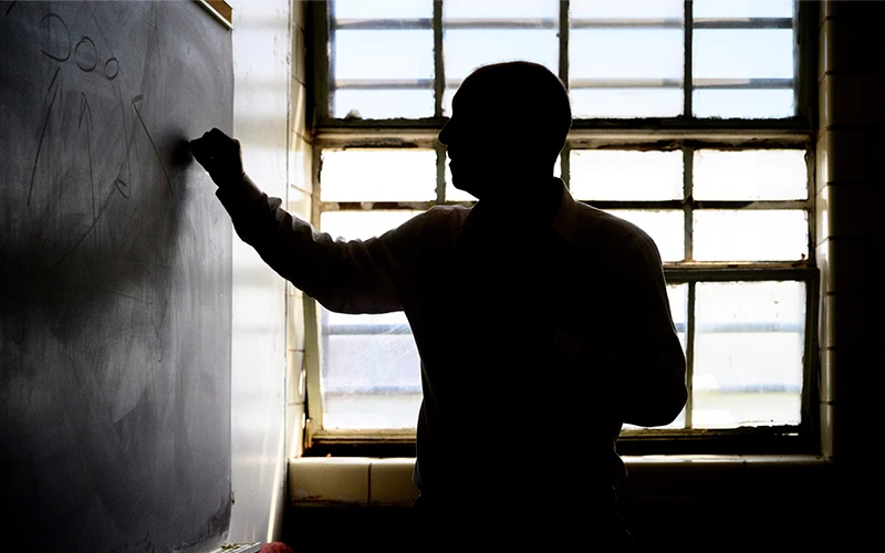 A man stands in profile writing on a chalkboard. He is silhouetted against a bright window.