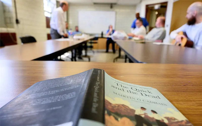 A close up of an open book face down on a table with people blurred out in the background.