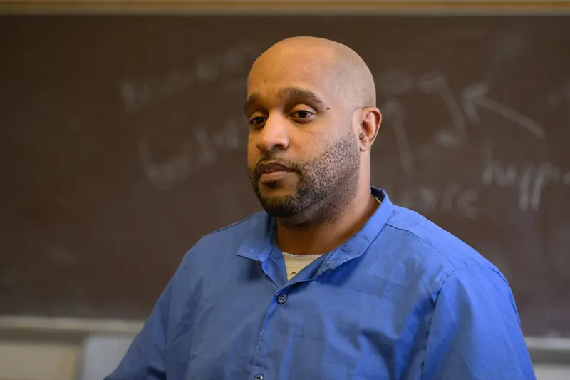 A man sitting in front of a chalkboard wearing a blue button-up shirt.