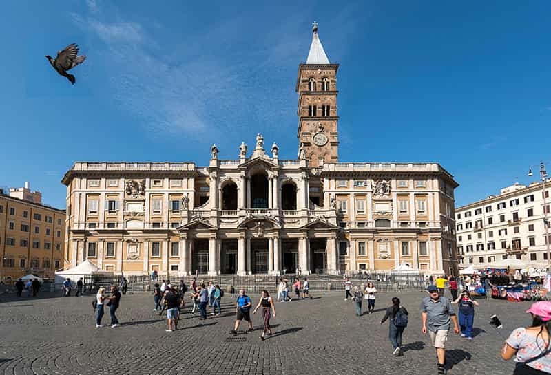 Tourists walk and take pictures in front of the Santa Maria Maggiore church, a bird is flying past in the foreground.