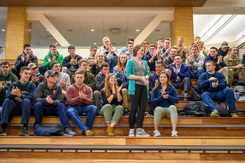 A student stands while other students sit and clap around her.