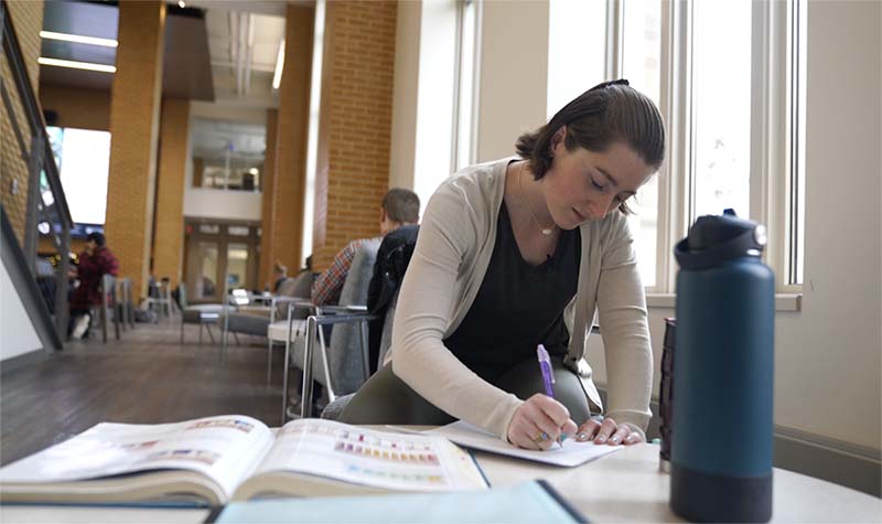 A female student sits at a table with an open textbook, taking notes.