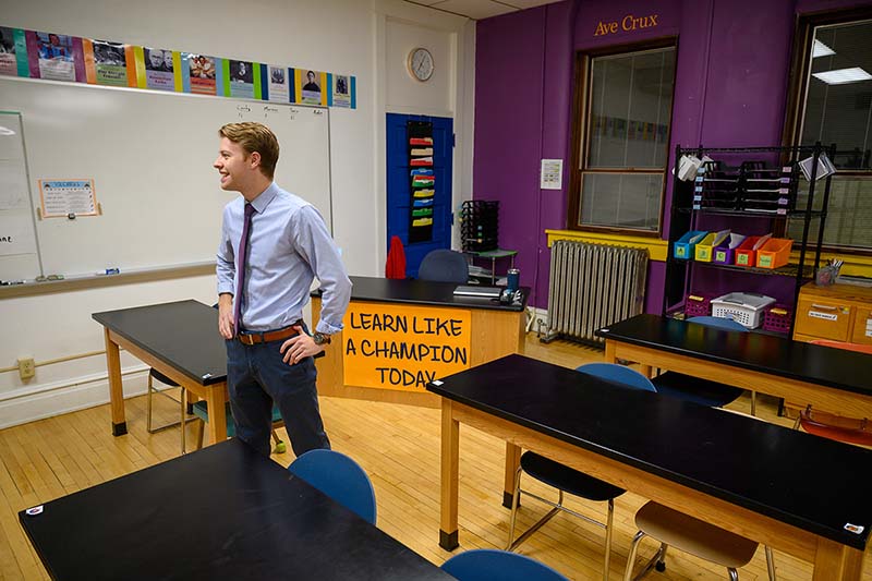 A smiling teacher stands in an empty classroom, with a sign that reads Learn Like a Champion Today hanginf from his desk.