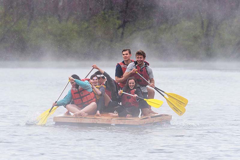Students on a homemade raft with oars on the lake on a cold rainy day.