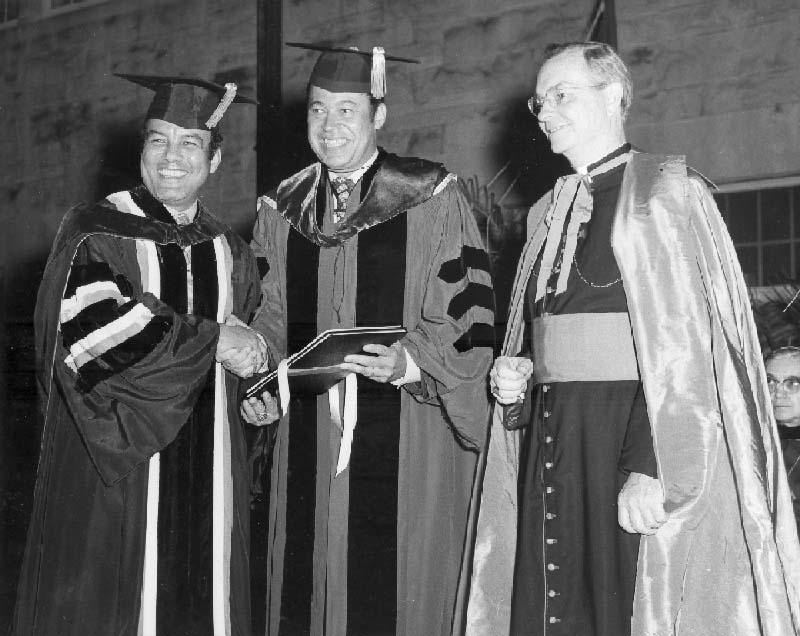 Black and white photo of Francis and Brooke in academic regalia shaking hands. A priest stands to the right of them.