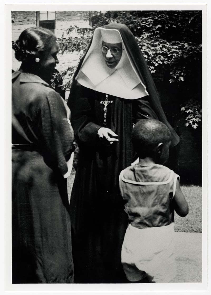 St. Katherine Drexel, dressed in nun attire with a crucifix around her neck, talks with a mother and child.