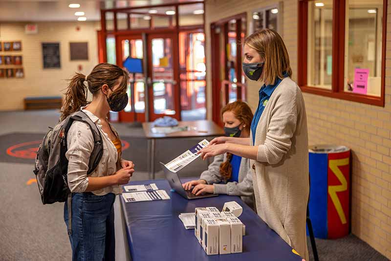 A girl wearing a backpack and mask speaks to	woman, also wearing a mask and pointing to a sheet of paper, about wifi hotspots.