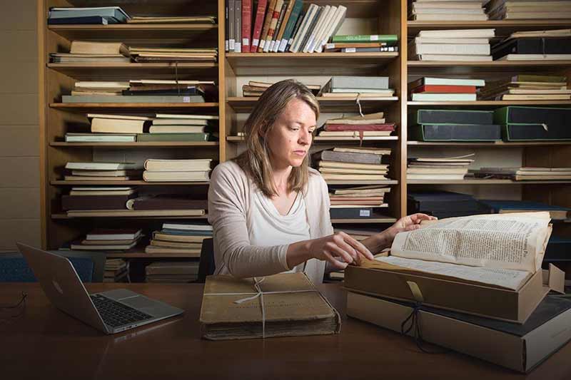 Elaine Stratton Hild at her desk, flips through an old book. Bookshelves line the wall behind her and her laptop is open on her desk.