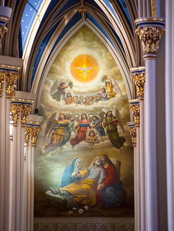 St. Joseph lies on a bed, with Mary and Jesus at his sides. A choir of angels hover over him, and a dove is above them.