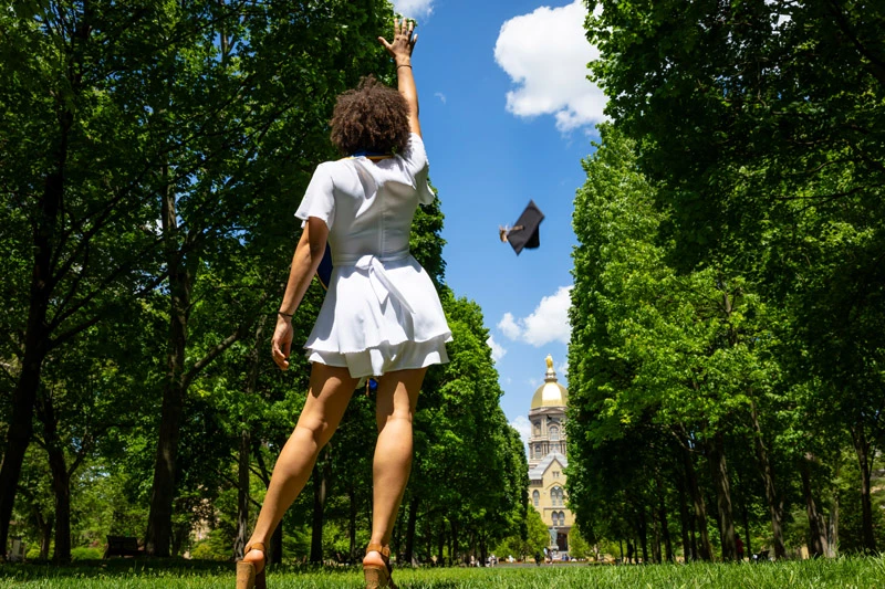 A graduating student in a white dress tosses their mortarboard into the air, with the Main Building's Golden Dome visible through the trees in the background.