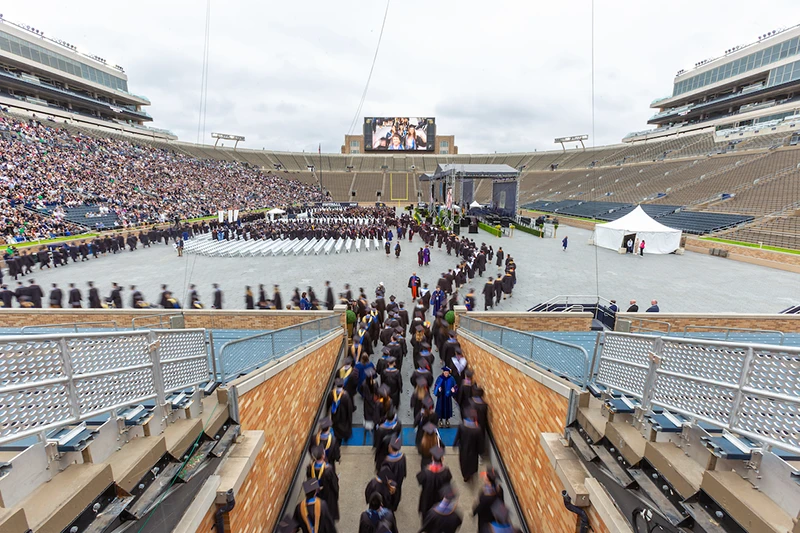 Graduates in black gowns and gold tassels process into Notre Dame Stadium for Commencement. The blurred motion shows them walking down ramps into the stadium, with spectators and rows of white chairs visible on the field.