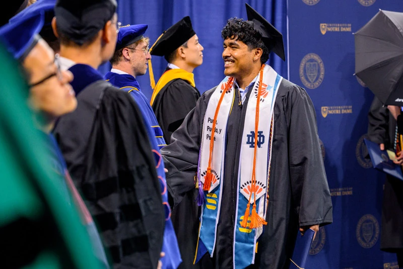 A smiling graduate in a black cap and gown with a white, orange, and teal stole smiles during the University of Notre Dame commencement ceremony.