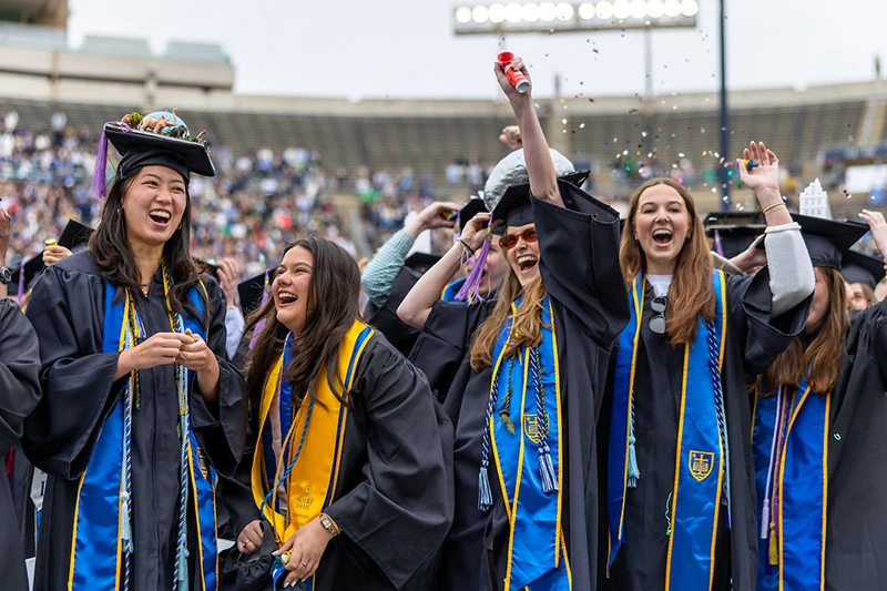 Notre Dame graduates in black gowns and blue and gold stoles celebrate with confetti and cheers in Notre Dame Stadium.