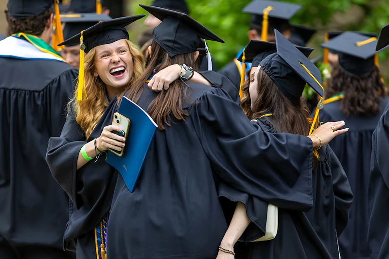 Three graduating students in black gowns and gold-tasseled mortarboards hug. One laughs, holding a phone and program.