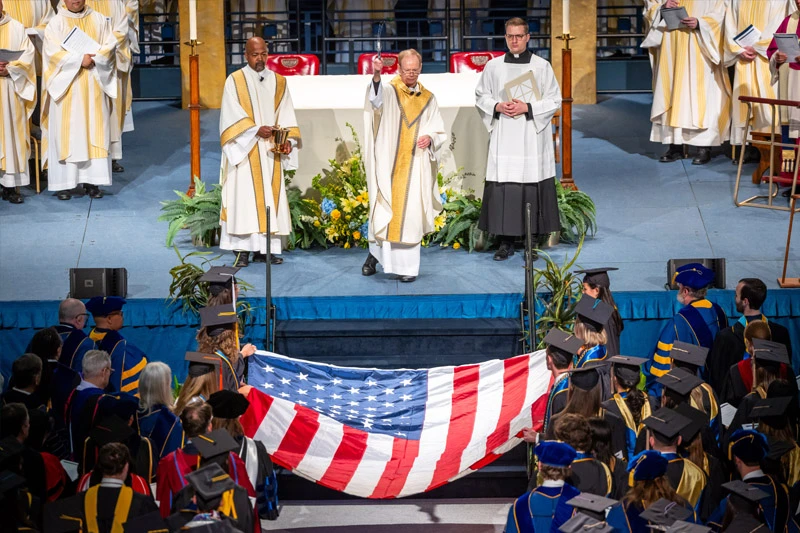 Graduates in academic regalia hold a large American flag at a ceremony, with clergy on a raised platform in the background.