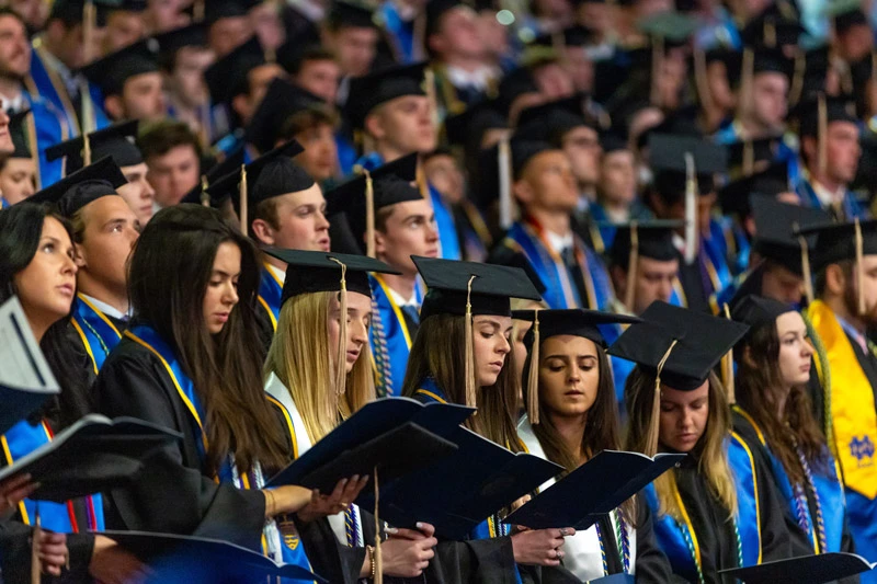 Notre Dame graduates in academic regalia with blue and gold stoles hold open books during the Commencement ceremony.