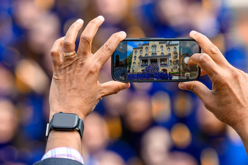 Hands hold a smartphone taking a photo of graduates in blue gowns gathered in front of Morrissey Hall at the University of Notre Dame.	A blurred crowd wearing blue is visible in the background.