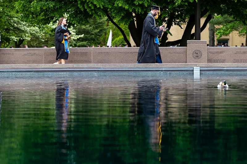 Two graduating students walk past the Clarke Memorial Fountain at the University of Notre Dame. One holds a program and the other looks at their phone. Their reflections are visible in the water, along with a duck.