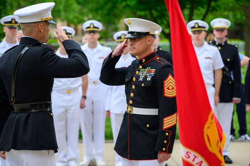 Two U.S. Marines in dress uniform salute each other beside a red Marine Corps flag.