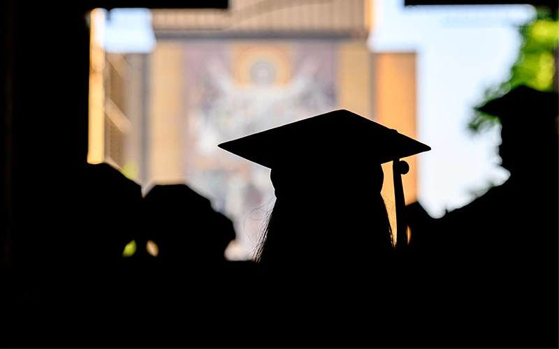 Silhouette of a graduating student wearing a mortarboard, with the Basilica of the Sacred Heart at Notre Dame blurred in the background.