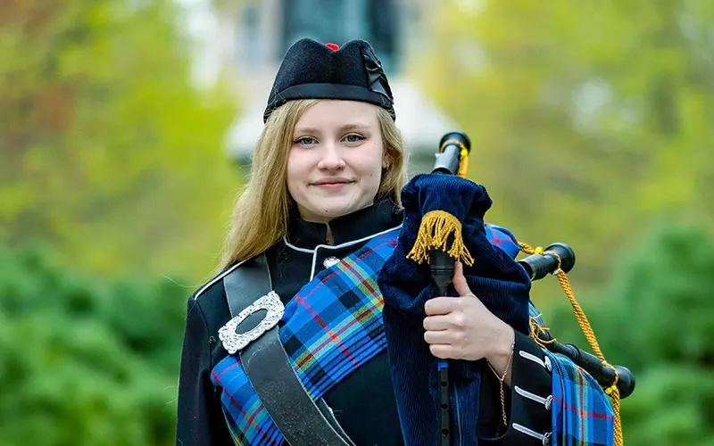Smiling blonde student in a black cap and dark uniform with a blue and green plaid sash holds bagpipes outdoors on a green, leafy campus.