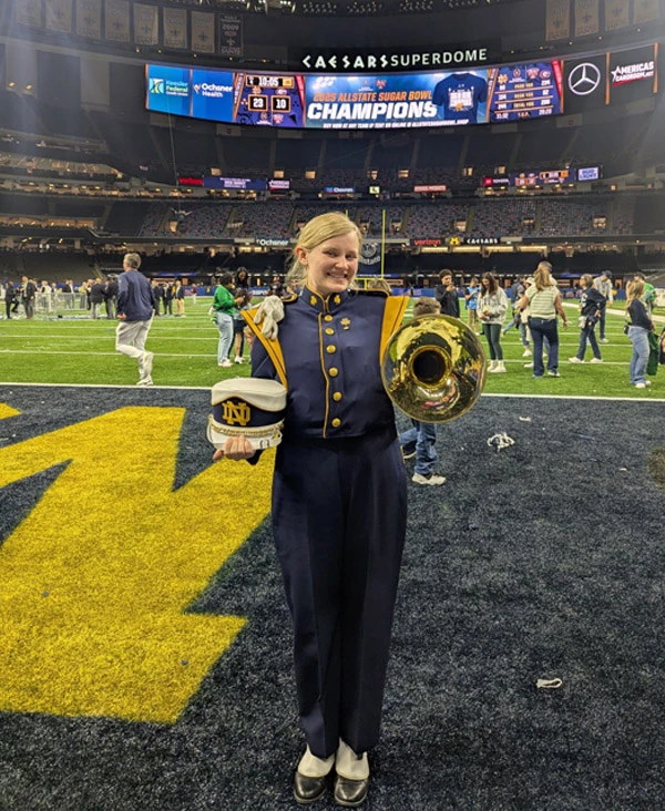 A smiling blonde female member of the Notre Dame Band holds her shako and mellophone on the field at the Caesars Superdome. A scoreboard displays the '2023 Allstate Sugar Bowl Champions' graphic.