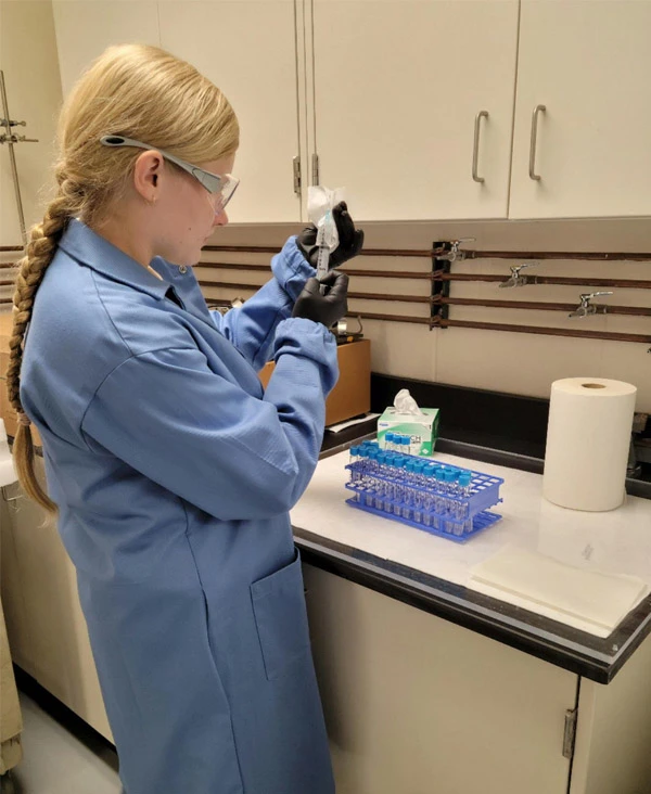 A white female with long blond hair wearing a blue lab coat, safety glasses, and black gloves prepares a syringe in a laboratory setting.	A rack of small, blue-capped test tubes sits on the counter beside them.