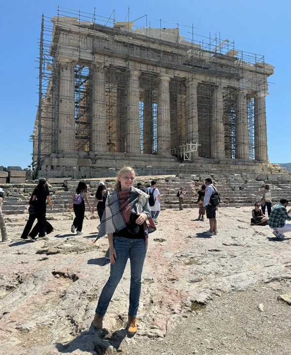 A blonde white female wearing jeans and a plaid shawl stands on the rocky ground in front of the Parthenon, partially covered in scaffolding, with other tourists in the background.