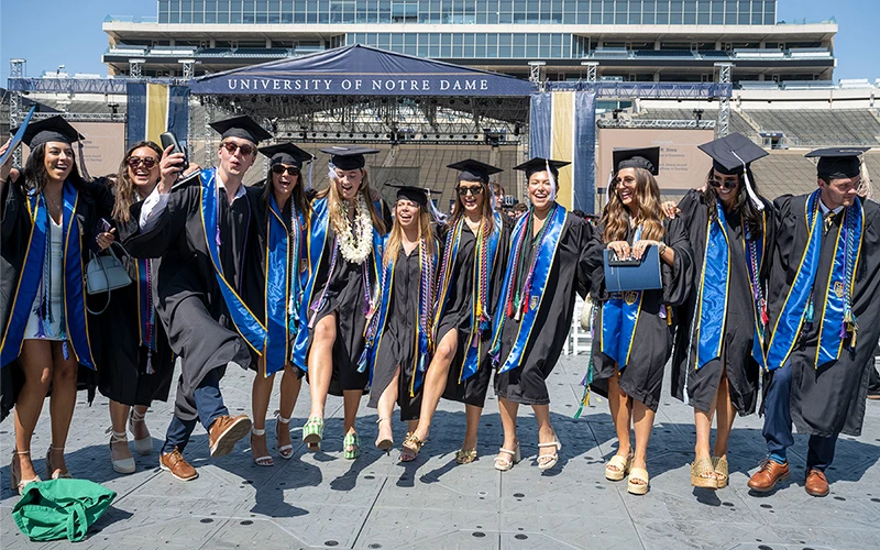 Graduates in black gowns and blue stoles celebrate with high kicks in front of the Notre Dame Stadium commencement stage.
