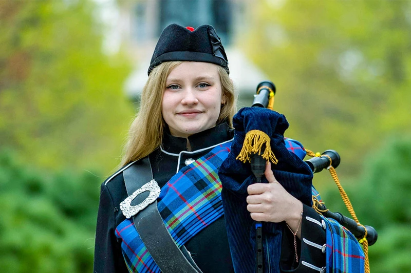 Smiling blonde student in a black cap and dark uniform with a blue and green plaid sash holds bagpipes outdoors on a green, leafy campus.