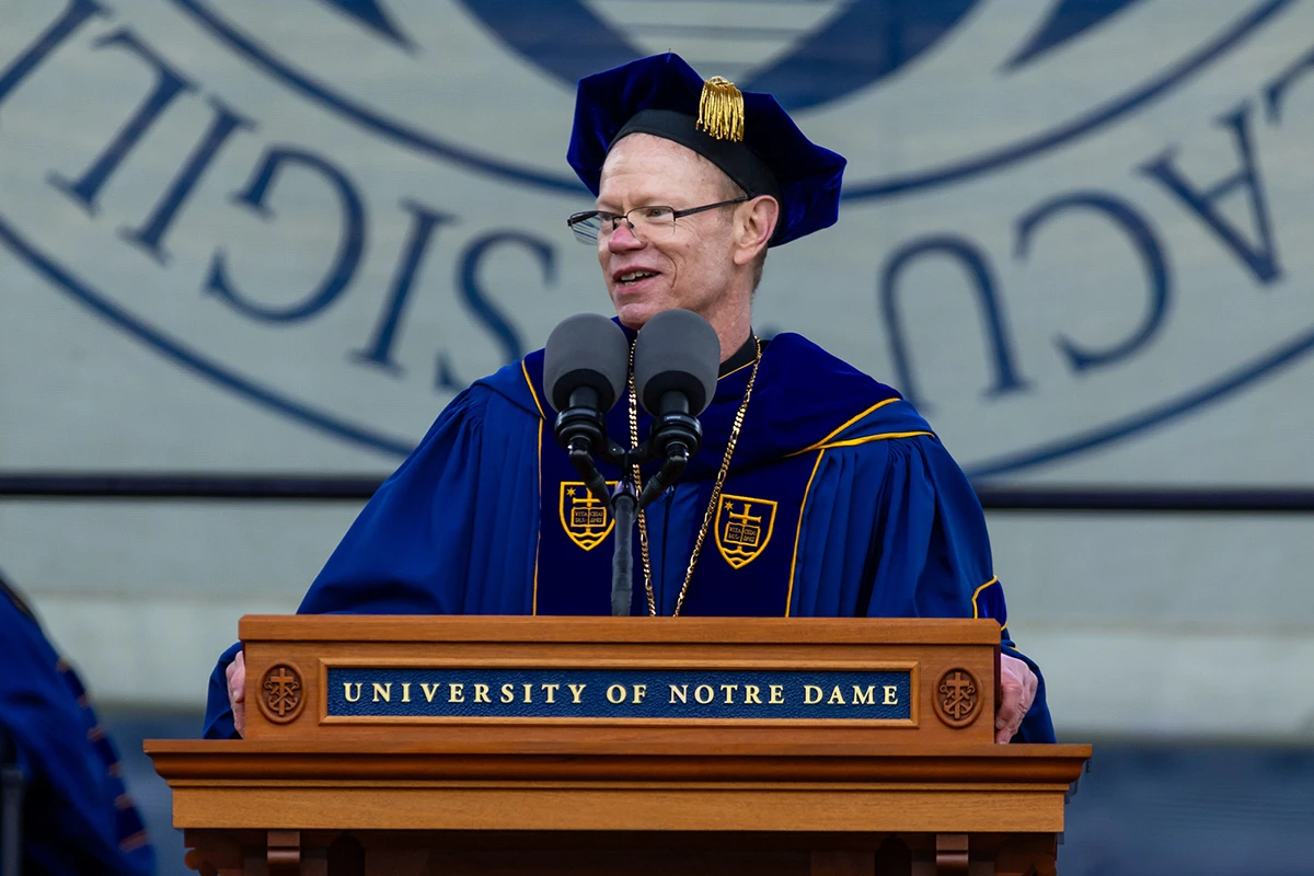 A person wearing a blue academic robe and mortarboard with gold accents speaks at a wooden podium bearing the University of Notre Dame logo.