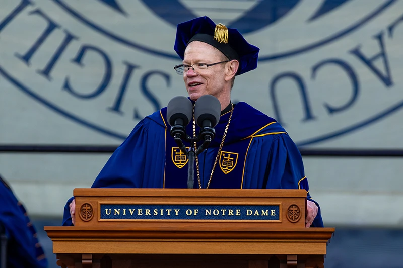 A man wearing a blue academic robe and mortarboard with gold accents speaks at a wooden podium bearing the University of Notre Dame logo.