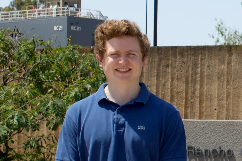 Smiling young man with curly red hair, wearing a blue polo shirt, stands in front of a tree and a sign for Notre Dame's Rancho Marino Reserve.