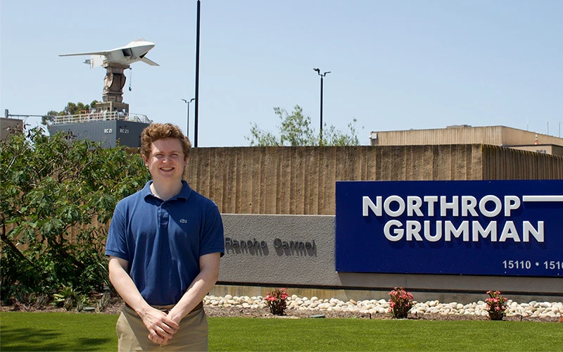 Red-haired man in a blue polo shirt and khaki pants smiles in front of a Northrop Grumman sign with a model B-2 Spirit stealth bomber in the background.