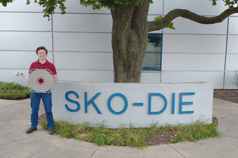 A person in a maroon shirt and jeans holds a large, octagonal, metallic object with a red circular center, standing in front of a sign reading 'SKO-DIE' outside a building.